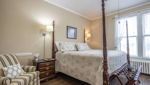 Una cama con dosel con cubrecama floral, una mesita de noche de madera con cajones, un sillón a rayas y un cuadro en la pared.
