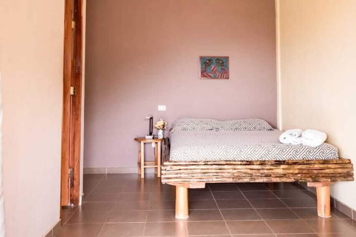 Un dormitori amb un llit, un ventilador de sostre, una taula petita amb una làmpada i un quadre a la paret.