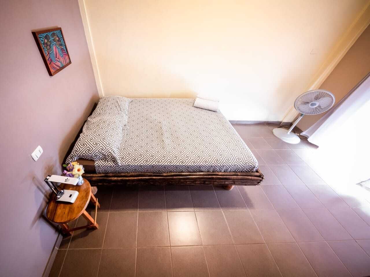 A compact room with a bed, a small table with a fan, and a wall-mounted picture.