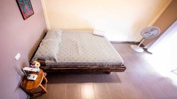 A compact room with a bed, a small table with a fan, and a wall-mounted picture.