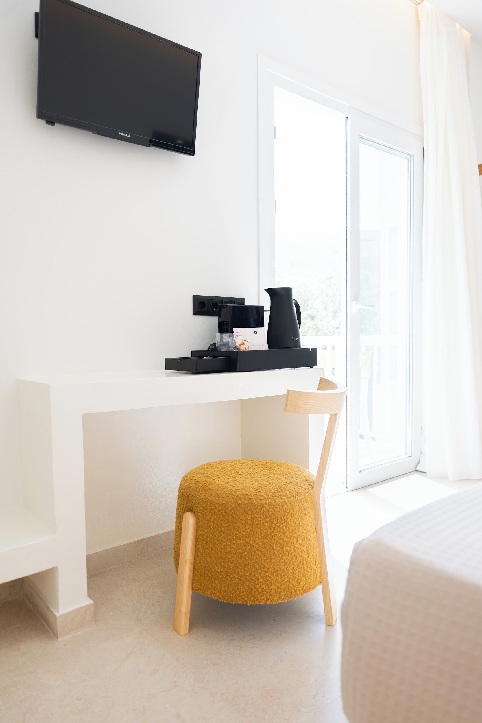 Habitación de hotel con un escritorio blanco, un televisor de pantalla plana montado, un taburete pequeño y una cama con cubierta blanca.