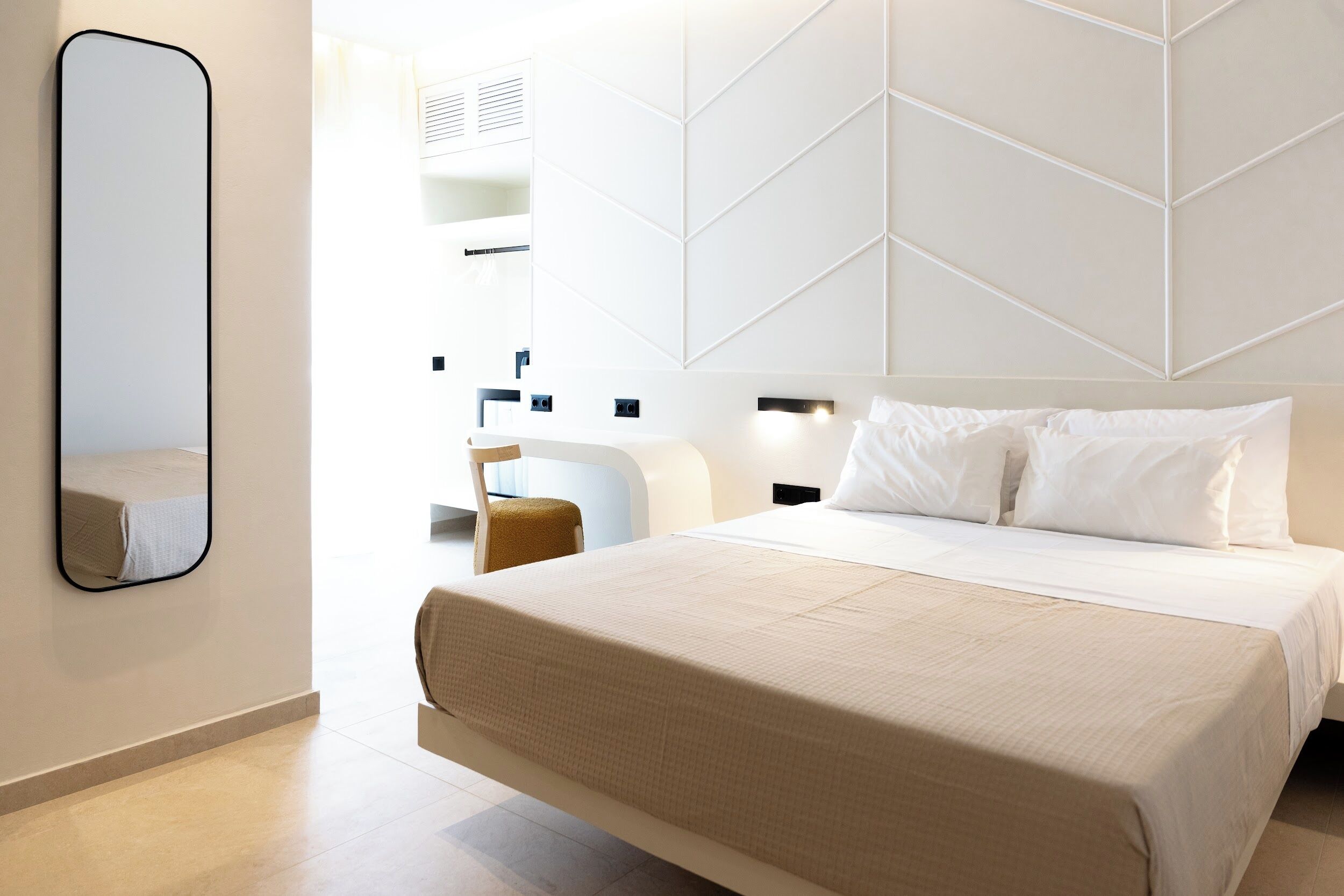 A modern hotel room with a large bed, a bedside table, a chair, and a mirror.