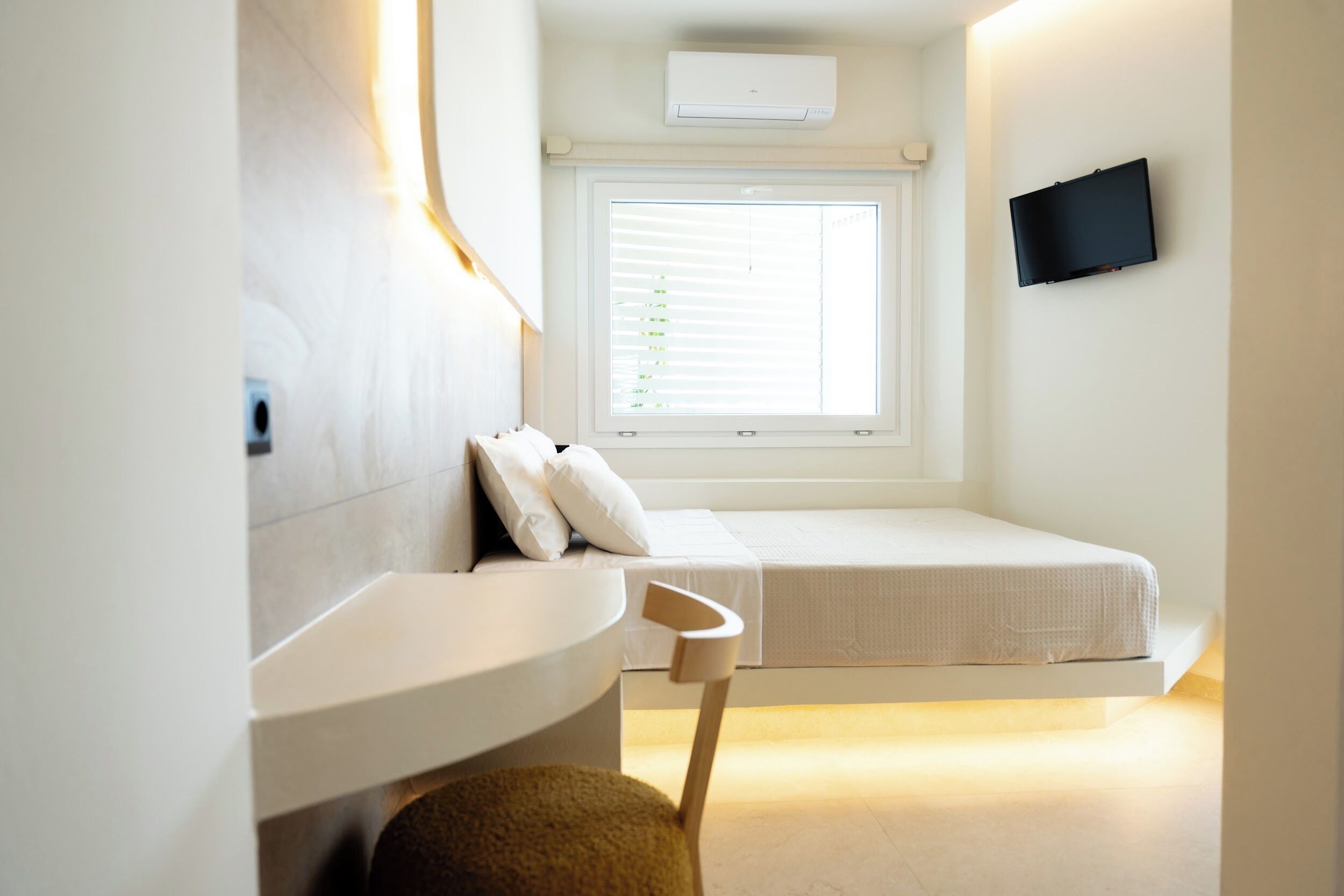 Un dormitorio pequeño con cama, mesita de noche, una silla, televisor montado en la pared y ventana con persianas.