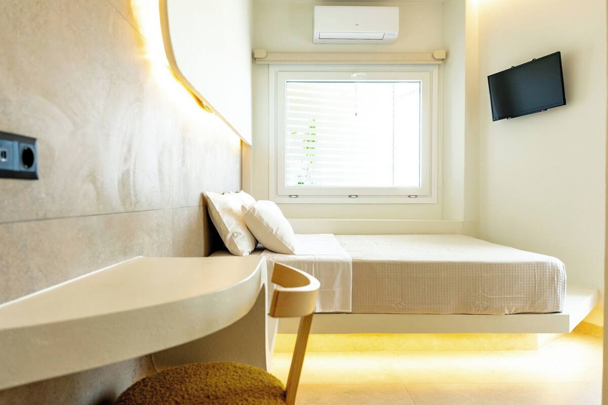 A modern room with a bed, a small table, a chair, a window with blinds, and a TV mounted on the wall.
