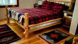 A wooden bed with a red and black checkered blanket, a wooden nightstand with a lamp, and a framed picture on the wall. - Mountain getaway in the heart of La Veta! (La Veta)