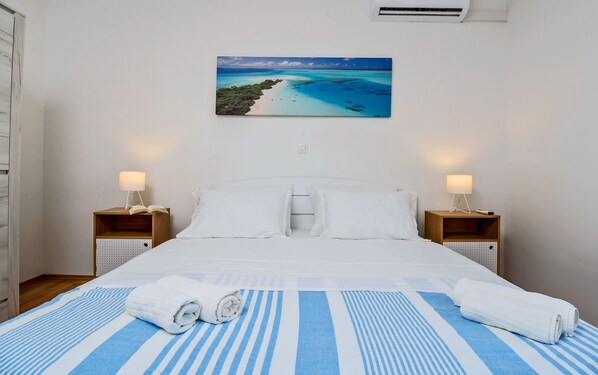 Una cama bien hecha con sábanas blancas, dos mesitas de noche con lámparas, un cuadro colgado en la pared de una playa y una ventana con un equipo de aire acondicionado.