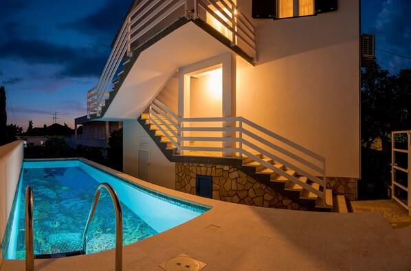A modern house with a swimming pool, a stone staircase, and a well-lit interior.