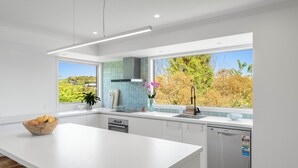 A modern kitchen with a large island, white cabinetry, a dishwasher, and a view of trees and houses through the windows.
