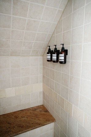 Un baño con baldosas color crema, un banco de madera y tres botellas de jabón líquido fijadas a la pared.
