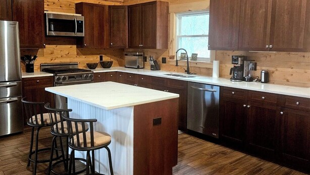 Una cocina amplia con armarios de madera, una isla central con taburetes de bar, electrodomésticos de acero inoxidable y un reloj en la pared.