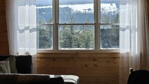 Interior de una cabaña con un letrero que dice 'Bienvenido a nuestra cabaña - La familia Neumann', una ventana grande con vistas a un paisaje nevado y una zona de estar acogedora con un sofá y sillones.