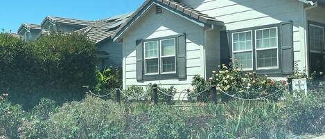 A two-story house with a gabled roof and shutters, surrounded by a garden with various plants and flowers.