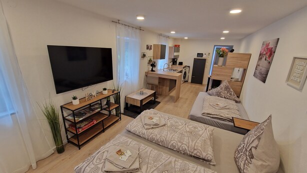 Un salon moderne avec un téléviseur à écran plat, un meuble TV en bois, un canapé, une petite table et un coin cuisine à l’arrière-plan.