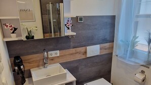 Une salle de bain moderne avec un lavabo blanc, des toilettes et un espace douche. Un miroir est placé au-dessus du lavabo, et une petite étagère y est fixée, sur laquelle sont disposés des objets décoratifs.