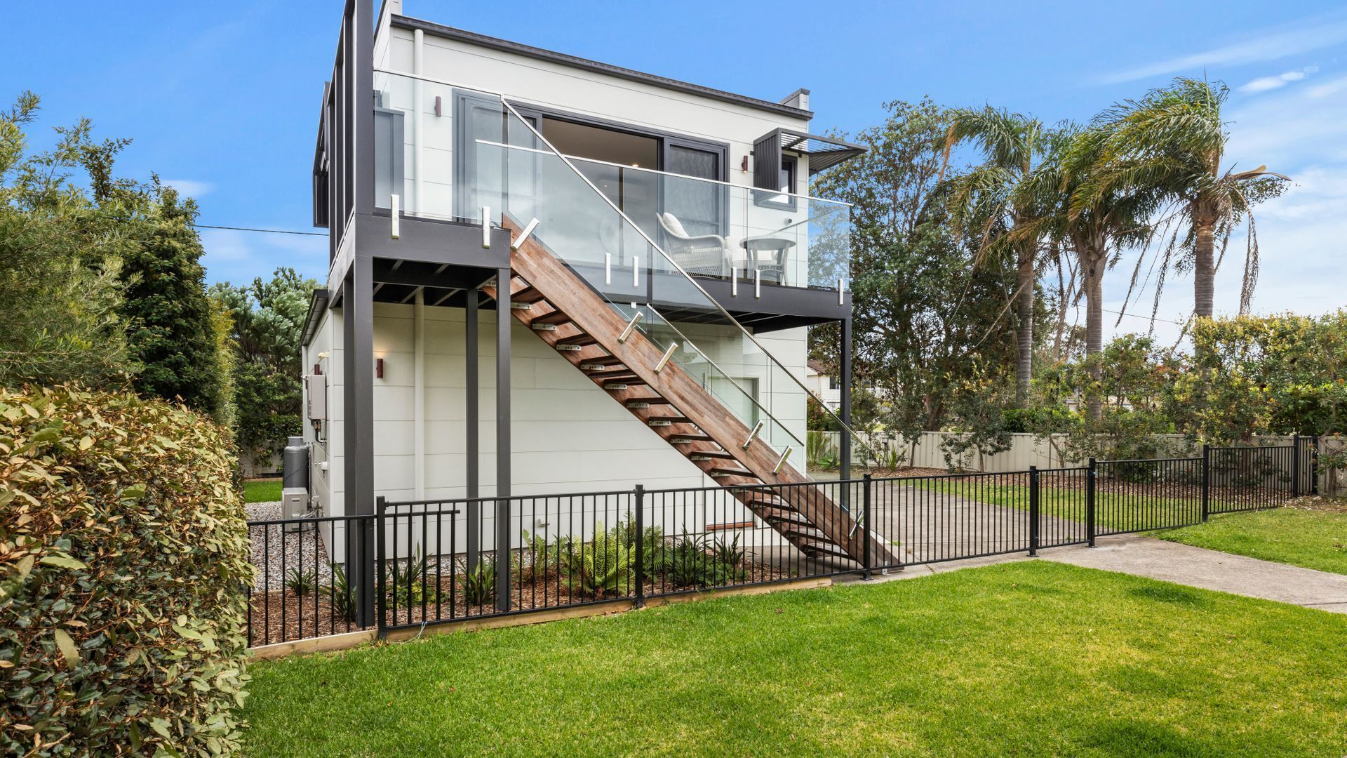 A modern two-story house with a glass balcony, a wooden staircase, and a fenced yard.