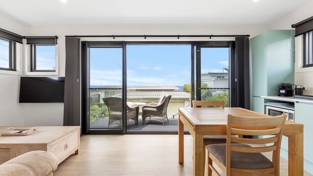 A modern kitchen with a built-in oven and a coffee machine, a dining area with a wooden table and chairs, and a sliding glass door leading to an outdoor patio with a view of the city. - Azure Studio - Ocean Glimpses - 3 Mins Walk to Beach (Callala Beach)