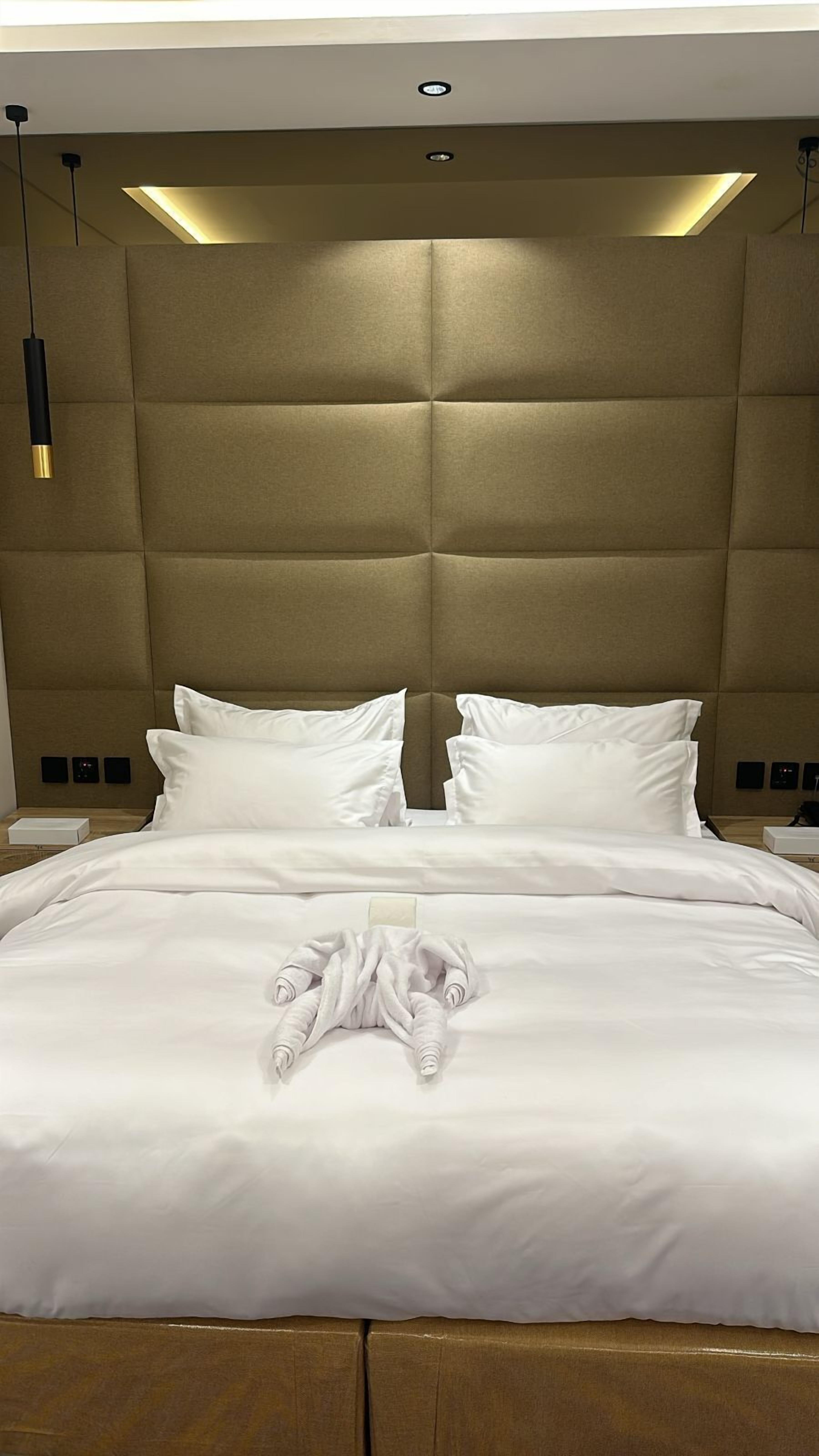 A modern hotel room with a large bed, a headboard with built-in lighting, and a bedside table with a lamp.
