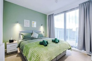Un dormitorio con cama, mesita de noche, ventana con cortinas y una obra de arte en la pared.