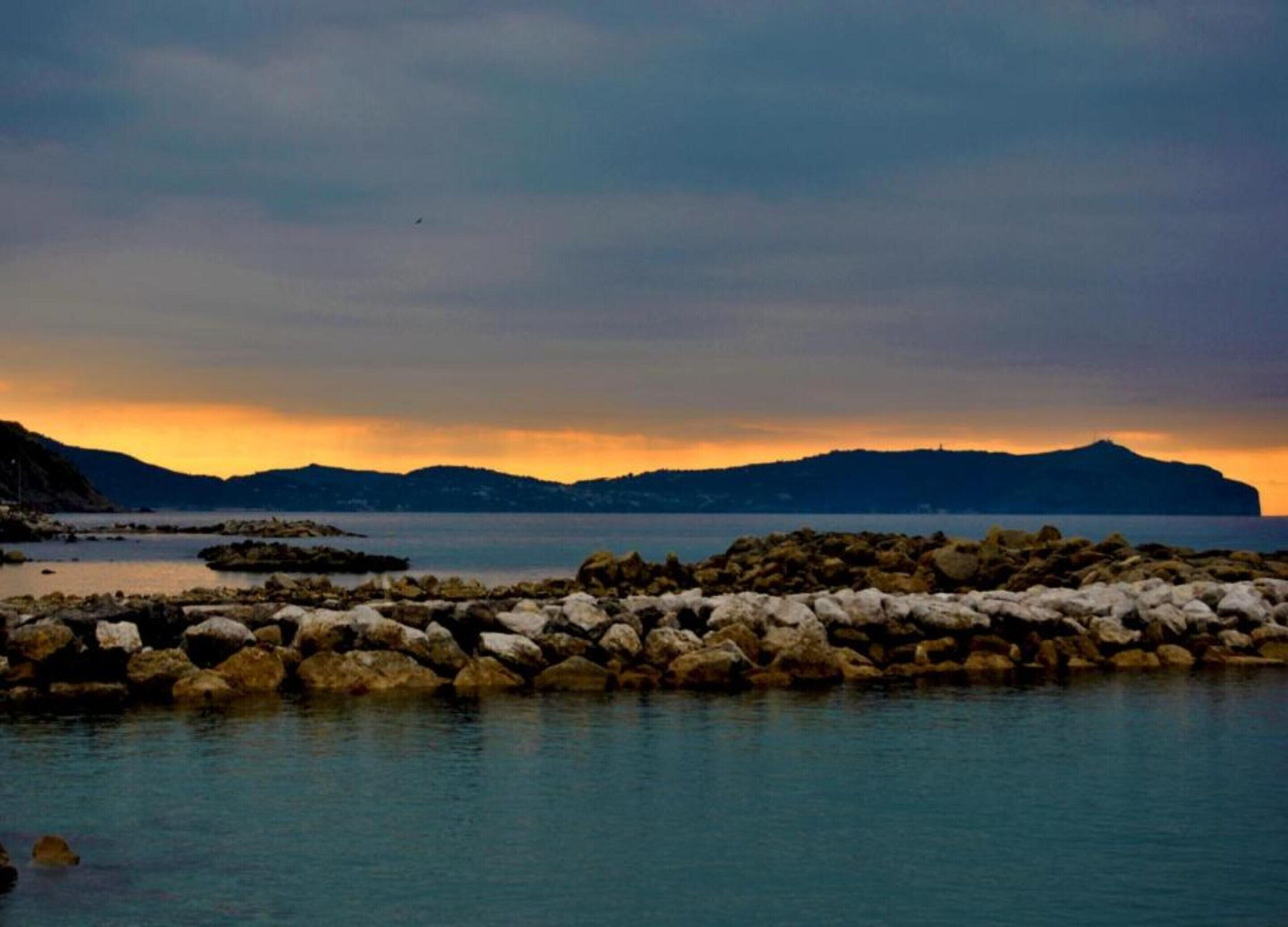 A coastal scene with a rocky shoreline, calm water, and distant mountains under a sunset sky.