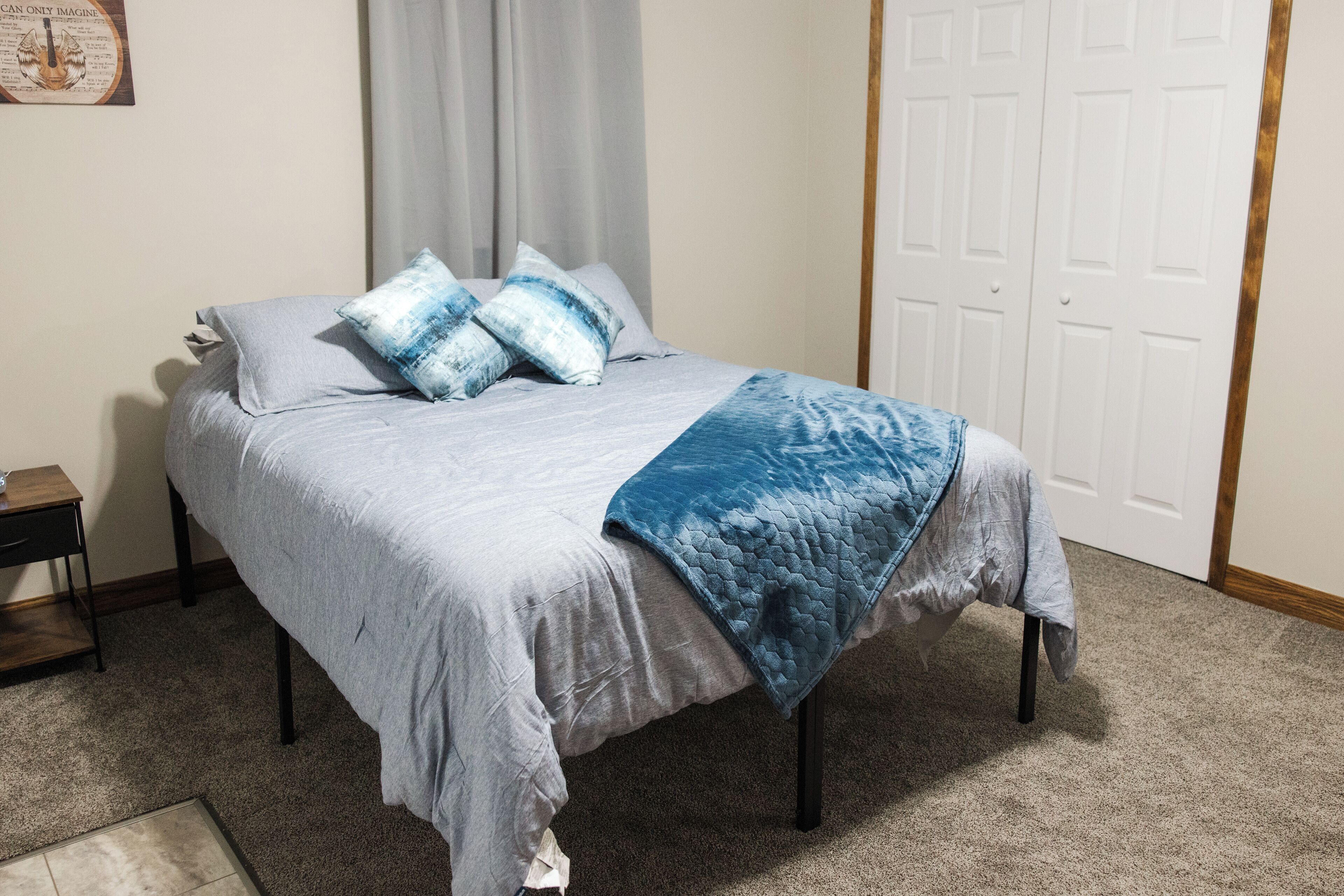A neatly made bed with a grey bedspread and blue pillows, a wooden nightstand with a lamp, and a white door in the background.