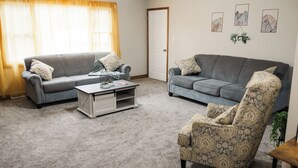 Ein Wohnzimmer mit zwei grauen Sofas, einem gemusterten Sessel, einem Couchtisch und einer Tür.