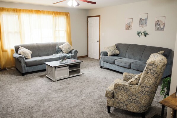 Ein Wohnzimmer mit zwei grauen Sofas, einem gemusterten Sessel, einem Couchtisch und einer Tür.