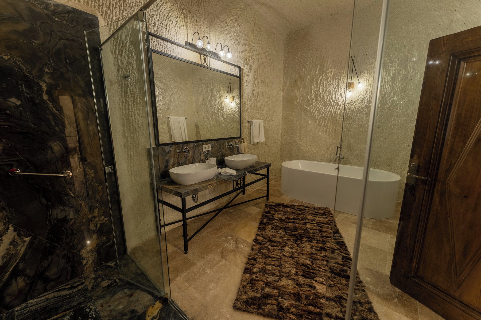 A bathroom with a glass shower enclosure, a large mirror, two sinks, a bathtub, and a wooden door.