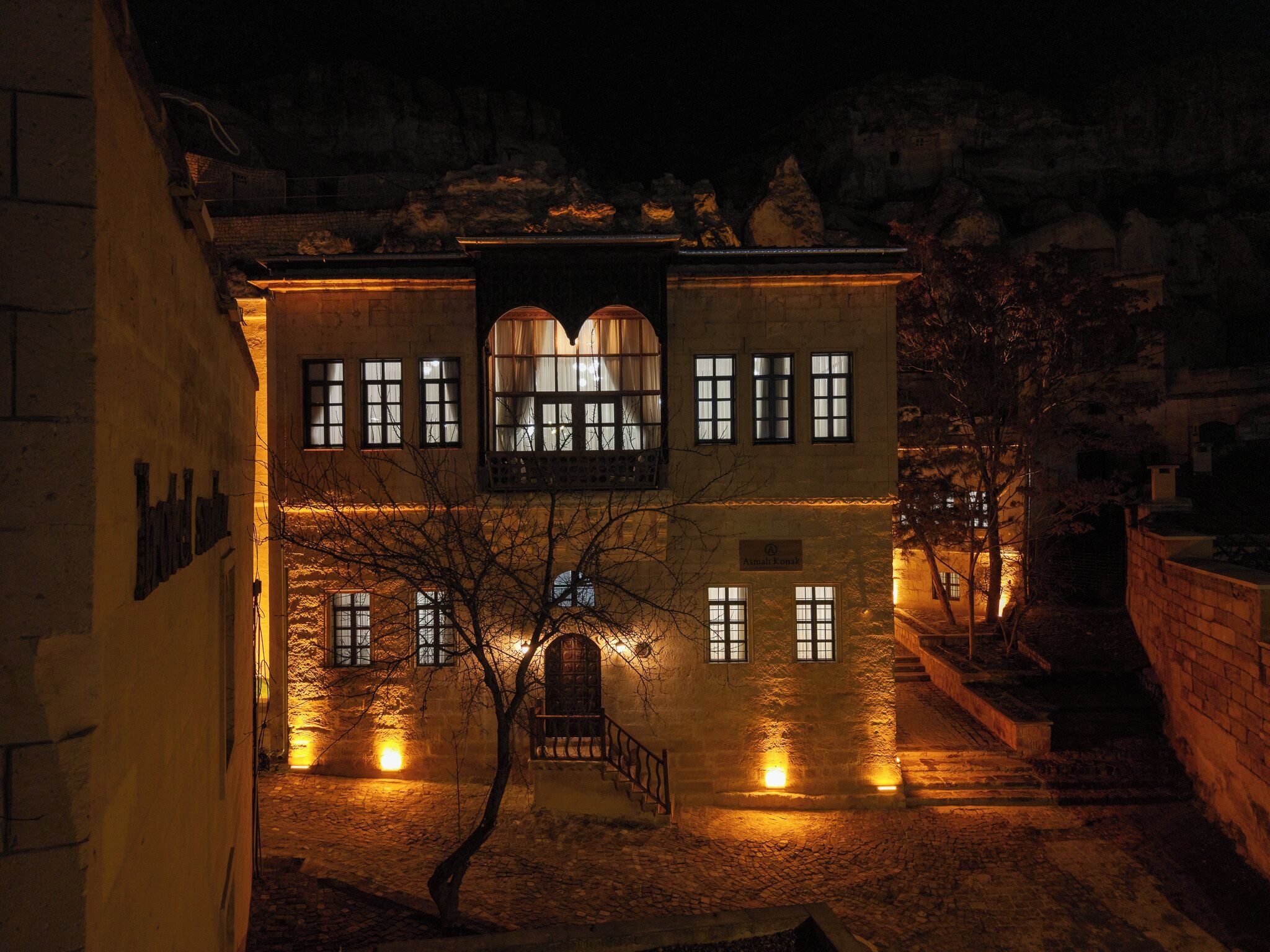 A historic building with multiple windows and a central arched entrance, surrounded by cobblestone pathways and a tree.