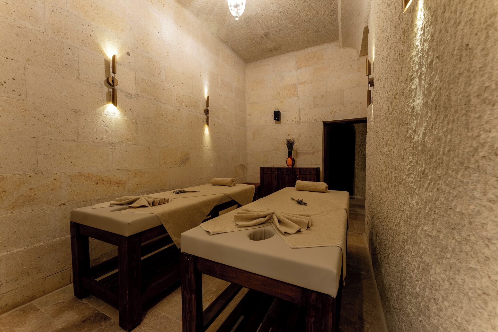 A spa room with two massage tables, a wooden bench, and a stone wall.