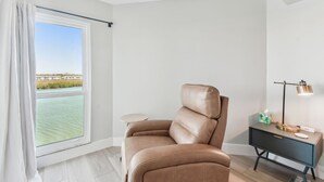 Ein modernes Zimmer mit Ledersessel, einem kleinen runden Tisch, einem Nachttisch mit Lampe und Blick auf ein Gewässer durch ein großes Fenster.