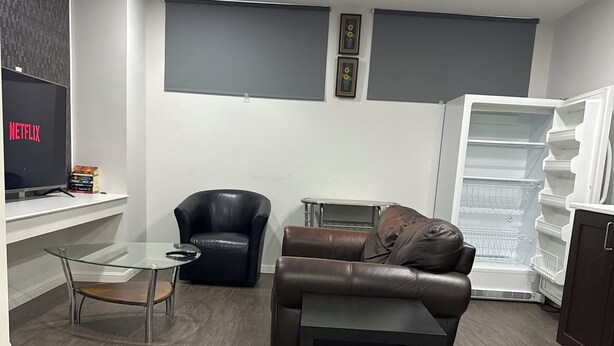 A modern living room with a leather sofa, a glass coffee table, a black leather chair, a TV displaying 'NETFLIX', and a white refrigerator. - The Last Resort with private entrance. In Basement. (Winnipeg)