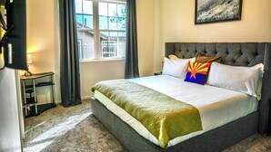 Una habitación de hotel con una cama grande, cabecero, una ventana con cortinas, una mesita de noche pequeña y un cuadro colgado en la pared.