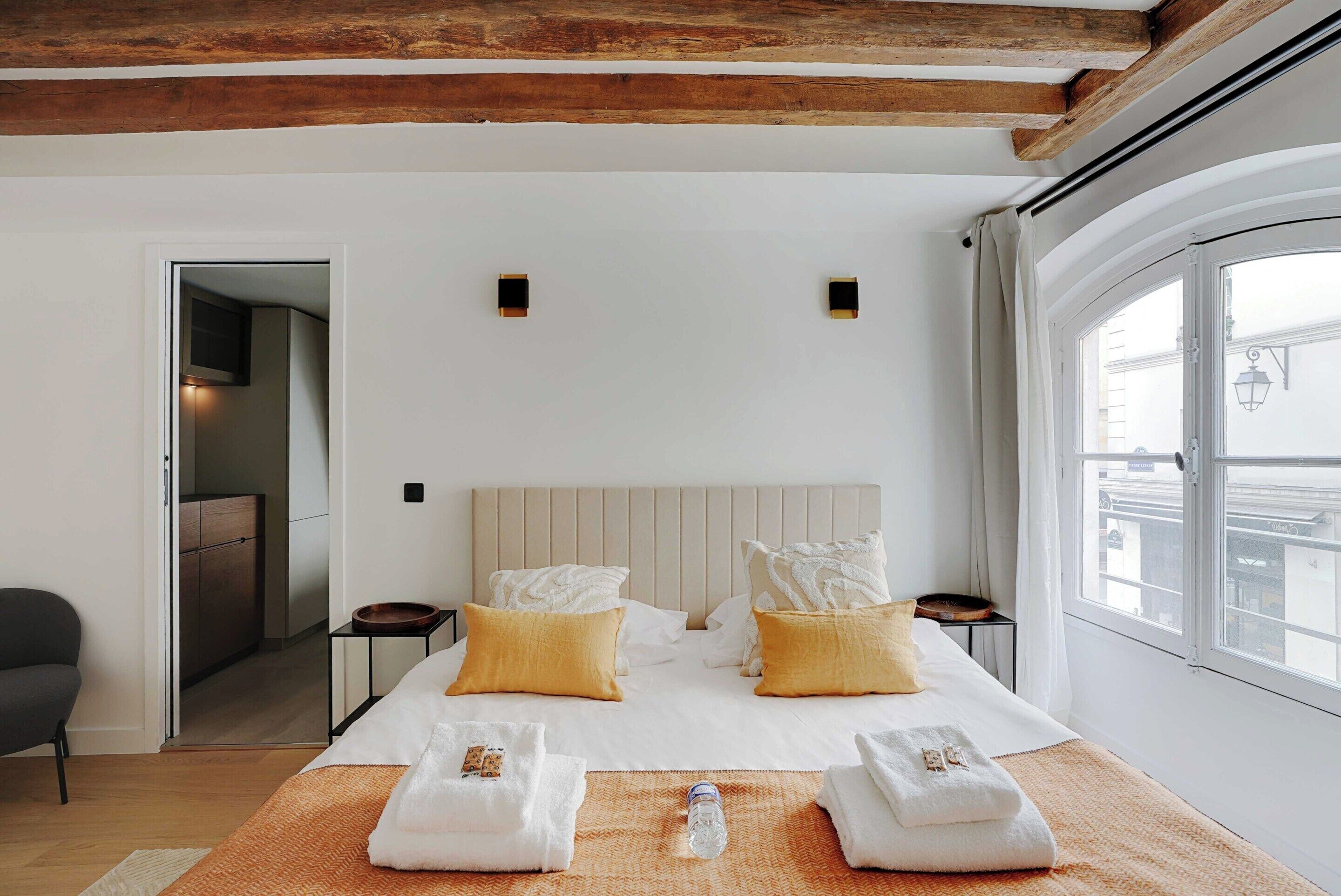 A hotel room with a large bed, two bedside tables, a window with curtains, and a wooden ceiling beam.