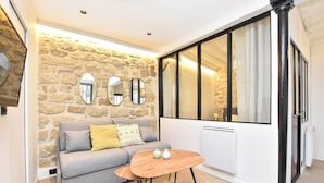 A modern living room with a stone wall, grey sofa, wooden coffee table, and large windows with black frames. - Cozy Apartment - 1br/4p - Sentier (Paris)