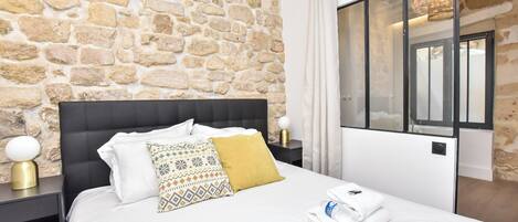 A hotel room with a stone wall, a black upholstered headboard, white bedding, a patterned throw pillow, a yellow accent pillow, a bedside table with a lamp, and a window with curtains.