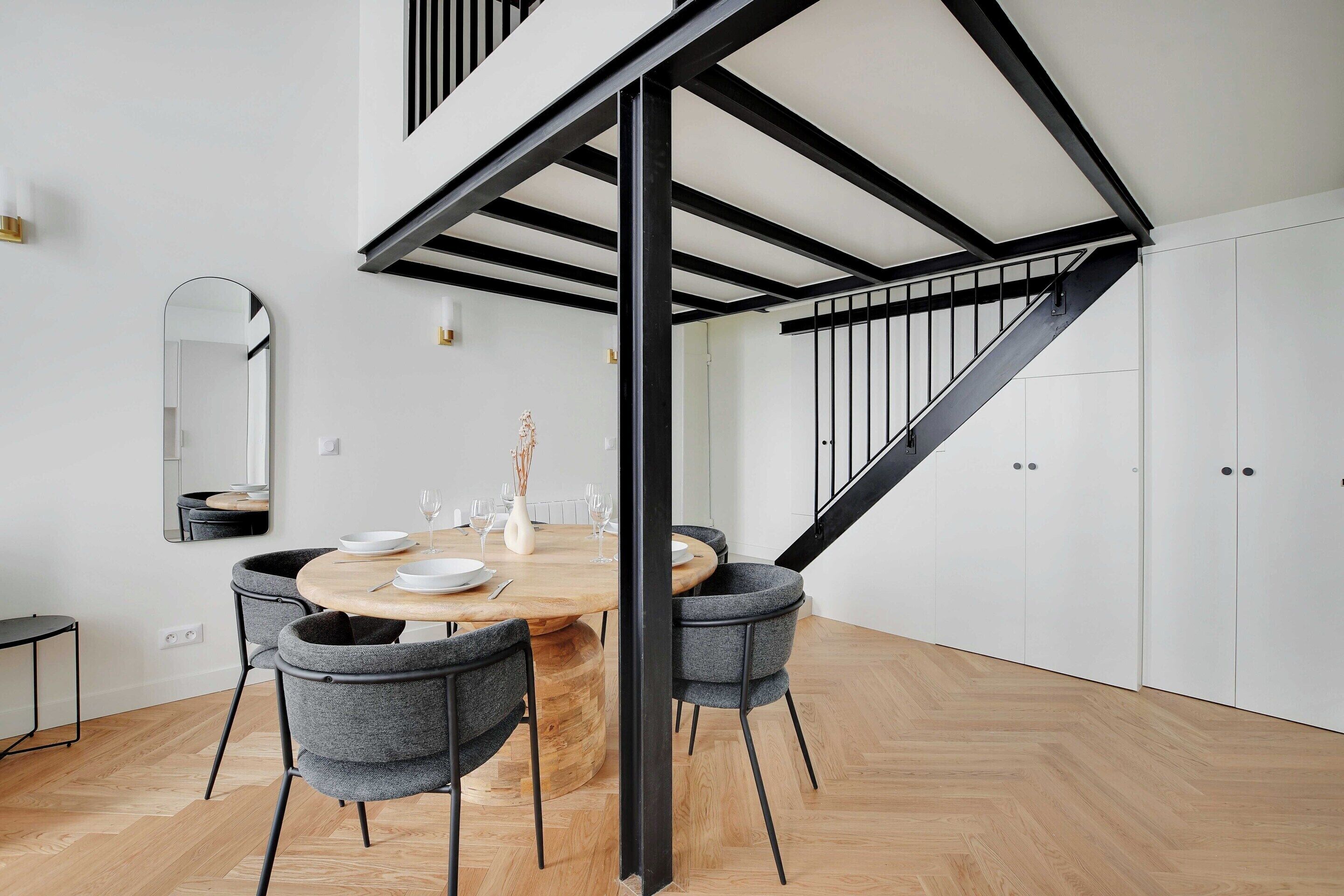 Un comedor con una mesa de madera y sillas grises tapizadas, una escalera negra y paredes blancas.