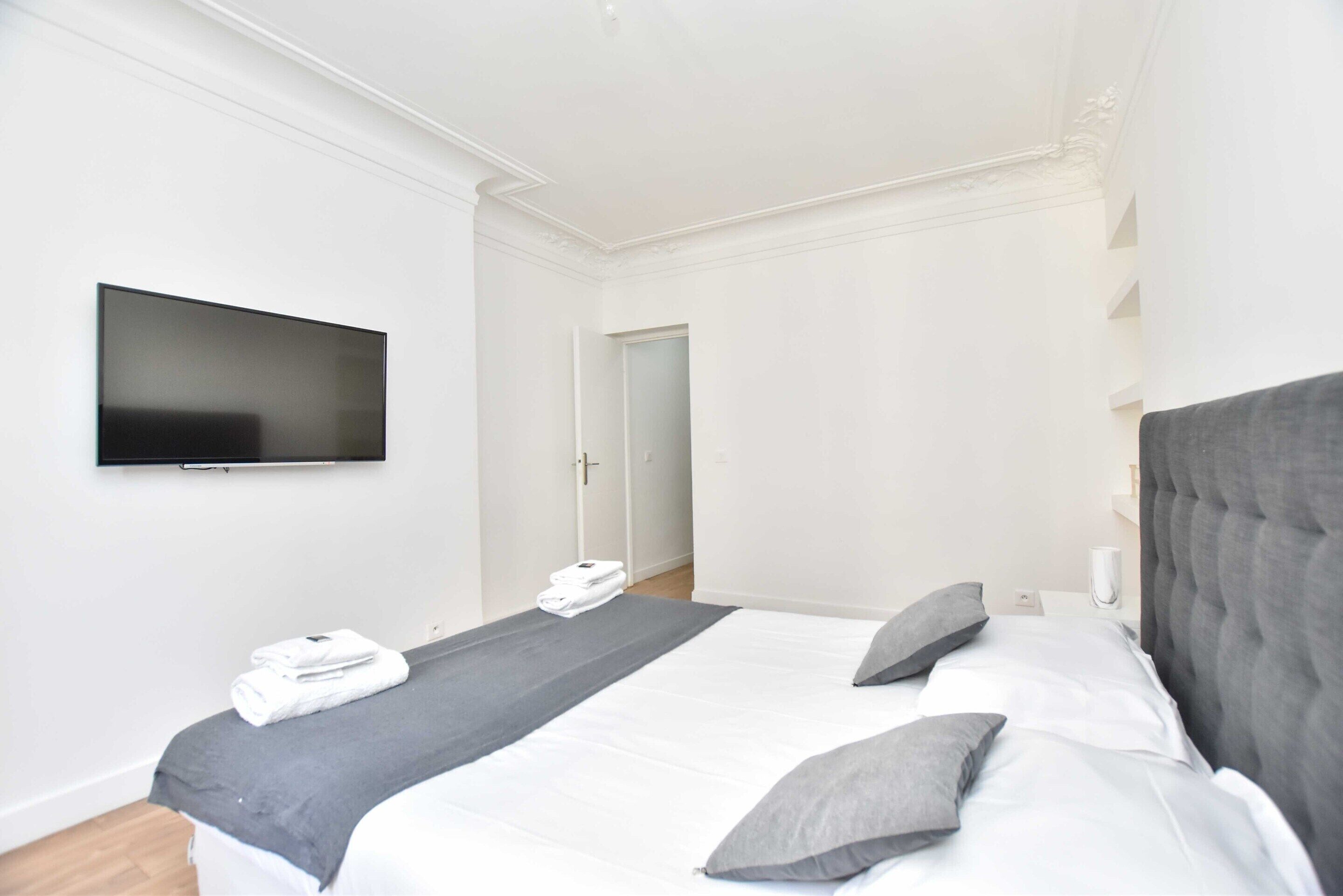 Una habitación de hotel moderna con una cama grande, un televisor de pantalla plana montado en la pared y una puerta que da a otra habitación.