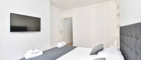 A modern hotel room with a large bed, a flat-screen TV mounted on the wall, and a door leading to another room.
