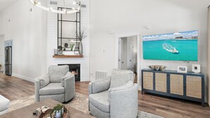 A modern living room with a fireplace, two grey armchairs, a coffee table with decorative items, a large flat-screen TV, and a wicker console. - Pools*Bikes*Pools*Walk2beach (Santa Rosa Beach)