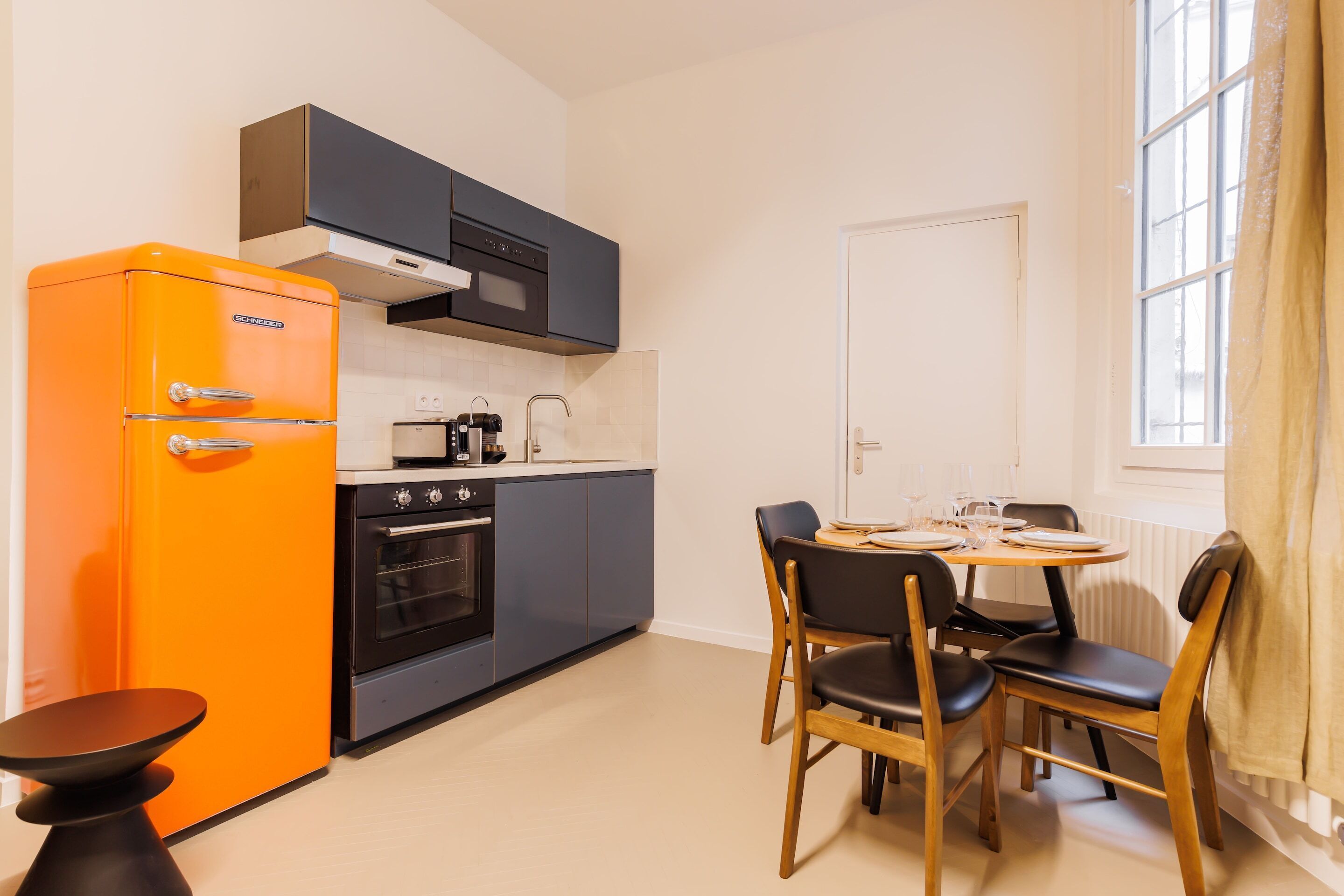 Eine kleine Küche mit einem altmodischen, orangefarbenen Kühlschrank, einem Herd und einer Mikrowelle. Ein Esstisch mit Stühlen und ein Fenster mit Vorhängen sind ebenfalls vorhanden.