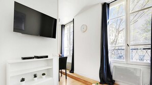 Una sala de estar moderna con un televisor de pantalla plana montado en la pared, un estante blanco con plantas decorativas, una silla negra y un ventanal con vistas a árboles y edificios.