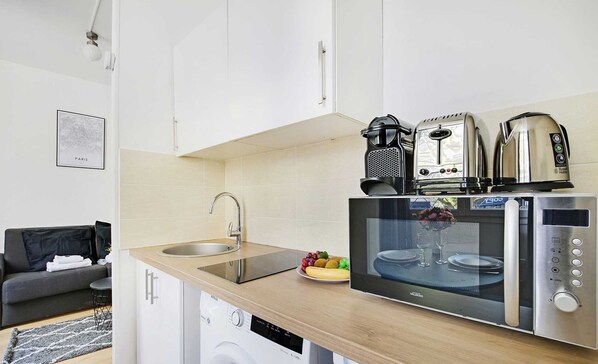 A compact kitchen with a sink, a microwave, a coffee maker, and a toaster. There is a washing machine below the counter, and a grey sofa in the background. - Le Marais - Rue de Bretagne (Paris)