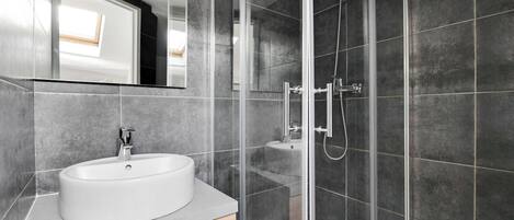 Un baño moderno con un lavamanos blanco, cabina de ducha con mampara de vidrio y paredes cubiertas de baldosas oscuras.