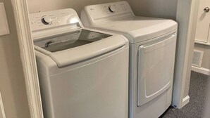 A laundry room with two white front-loading washers and dryers, a wooden shelf above, and a gray carpeted floor. - Down Home Comfort - Greenville, SC

 (Piedmont)