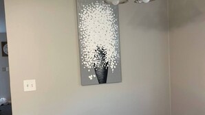 A dining area with a wooden table and chairs, a hanging light fixture, and a wall-mounted painting of a tree with white blossoms. - Down Home Comfort - Greenville, SC

 (Piedmont)