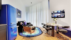 Una habitación de hotel moderna con un refrigerador azul, una zona de comedor con una mesa redonda y taburetes de bar, un televisor grande mostrando series internacionales y una ventana con cortinas blancas.