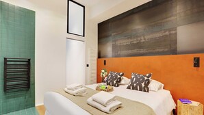 Habitación de hotel con una cama, una obra de arte colgada en la pared, una mesita de noche de madera y una zona de ducha con azulejos verdes.
