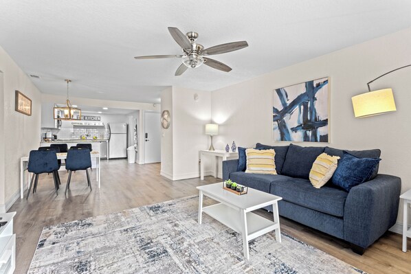 A modern living room with a grey sofa, white coffee table, and a dining area with chairs and a kitchen in the background. - Azure 2BR/2BA Condo , Heated Pool Near Siesta (Sarasota)