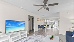 En moderne stue med et fladskærms-tv, en loftsvifte, et spiseområde med stole og et bord, og et køkken i baggrunden.