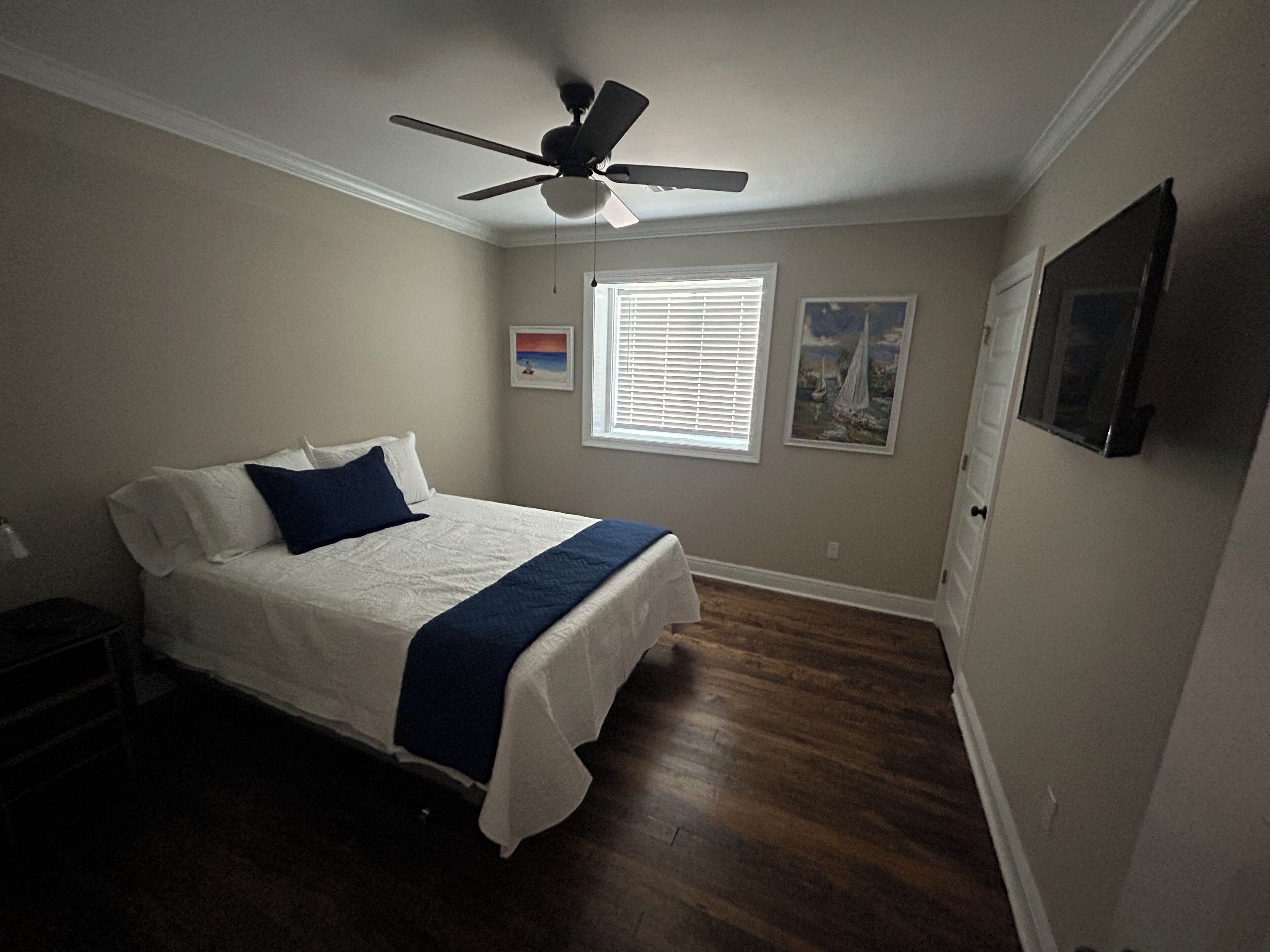 Une chambre à coucher avec un lit, un ventilateur de plafond, une fenêtre avec des stores et un tableau encadré au mur.